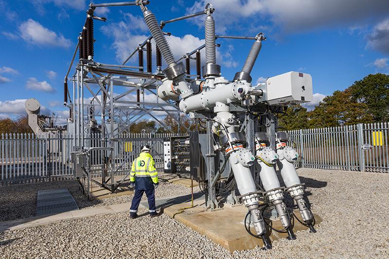 An electrical substation with wires and coils in a metal structure, 铺着砾石地板，周围有金属栅栏. 中间是戴着安全帽的G2能源公司职员