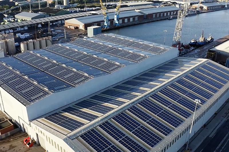 A view of a warehouse roof from a high elevation, with blue solar panels covering the roof. 仓库位于河边的工业区